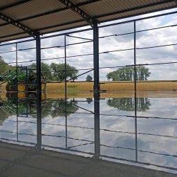 JUR-BUD Jakub Juraszek - Świeżo wylana, błyszcząca posadzka wewnątrz stalowej konstrukcji hali, odbijająca niebo i pole zboża widoczne na zewnątrz, z pracownikiem na podnośniku i traktorem w tle.