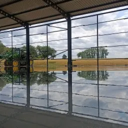 Świeżo wylana, błyszcząca posadzka wewnątrz stalowej konstrukcji hali, odbijająca niebo i pole zboża widoczne na zewnątrz, z pracownikiem na podnośniku i traktorem w tle.