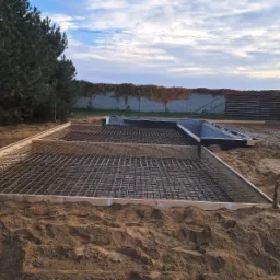 Przygotowany szalunek i siatka zbrojeniowa na piasku, gotowe do zalania fundamenty domu jednorodzinnego, w tle ogrodzenie z bluszczem.