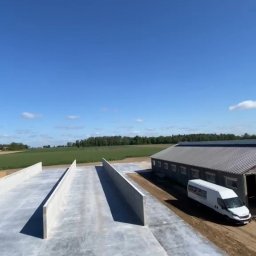 JUR-BUD Jakub Juraszek - Betonowa konstrukcja z wysokimi ścianami, prawdopodobnie fundamenty pod budynek gospodarczy, z widocznym budynkiem w tle i zaparkowanym białym busem.