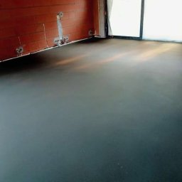 JUR-BUD Jakub Juraszek - Gładka, ciemna posadzka w surowym wnętrzu z ceglanymi ścianami i dużym oknem balkonowym. Widoczne ślady po instalacjach na ścianach.