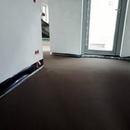 JUR-BUD Jakub Juraszek - Wnętrze w trakcie prac wykończeniowych: wylewka podłogowa, folia ochronna przy ścianach, widoczne gniazdka elektryczne. Okno balkonowe w tle.