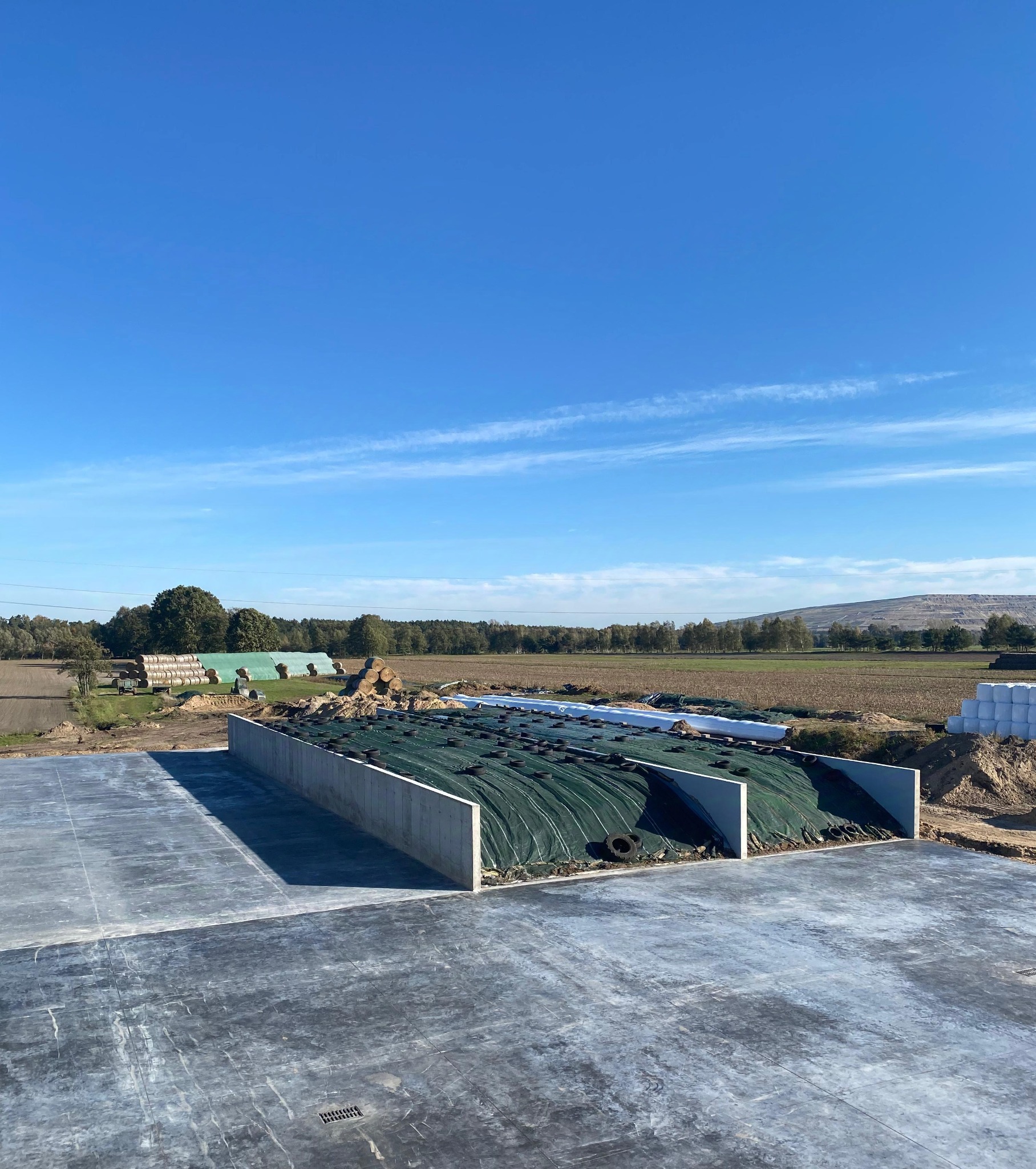 Widok z góry na betonową płytę i silos na kiszonkę z zieloną plandeką obciążoną oponami, w tle pola uprawne i błękitne niebo.