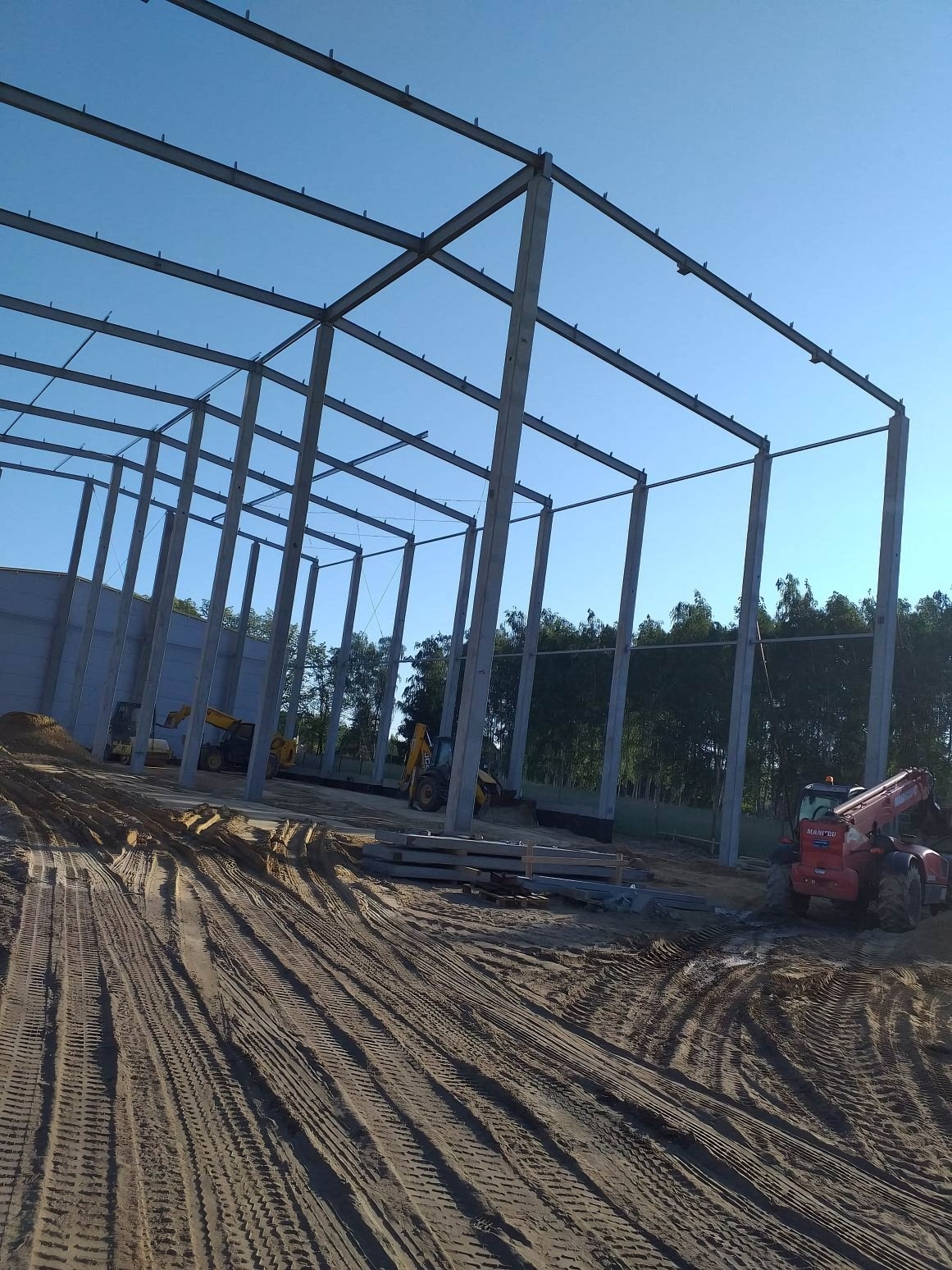 Wznoszenie konstrukcji stalowej hali z prefabrykatów żelbetowych, widoczne maszyny budowlane i świeżo wyrównany teren.