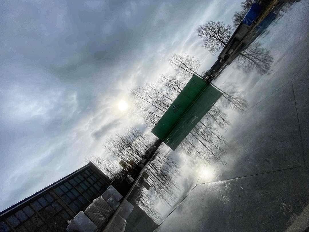 Błyszcząca, ciemna posadzka odbija pochmurne niebo, drzewa i fragment budynku, tworząc abstrakcyjne wrażenie głębi.