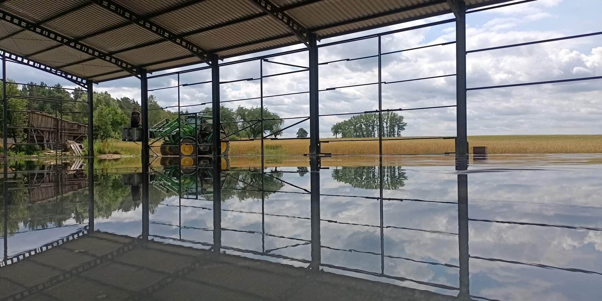 Świeżo wylana, błyszcząca posadzka wewnątrz stalowej konstrukcji hali, odbijająca niebo i pole zboża widoczne na zewnątrz, z pracownikiem na podnośniku i traktorem w tle.