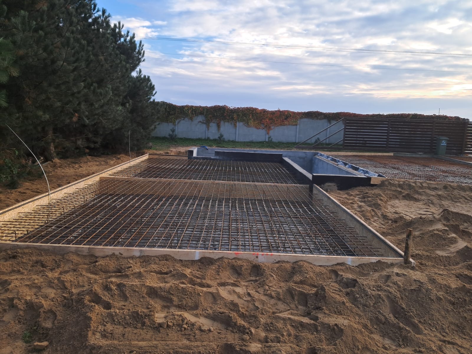 Przygotowany szalunek i siatka zbrojeniowa na piasku, gotowe do zalania fundamenty domu jednorodzinnego, w tle ogrodzenie z bluszczem.