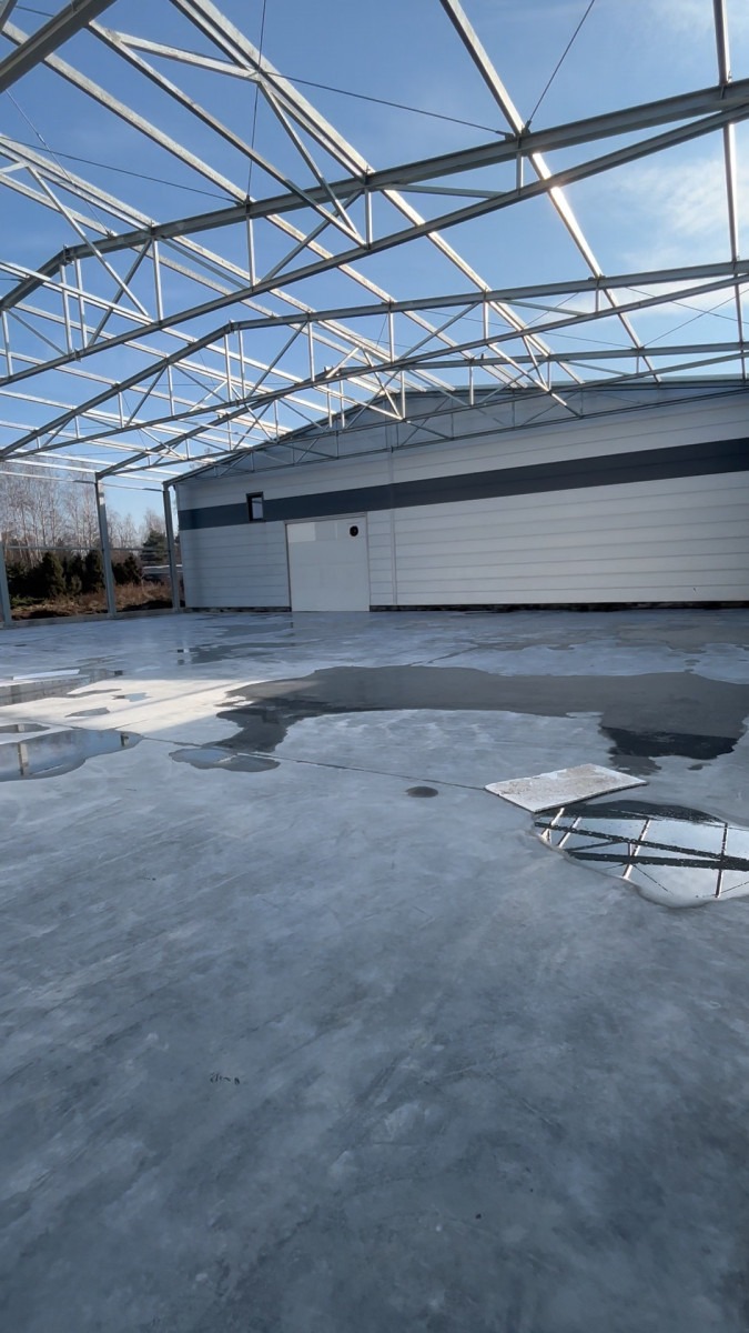 Wnętrze hali w budowie z widoczną stalową konstrukcją dachu i mokrą, betonową posadzką. W tle ściany z płyt warstwowych i drzwi.