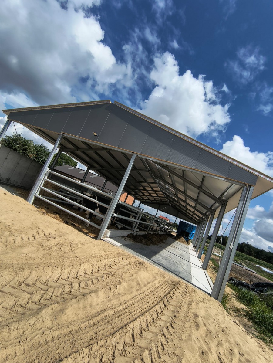 Zadaszona konstrukcja stalowa z szarym dachem, prawdopodobnie wiata dla zwierząt, widziana z zewnątrz na tle błękitnego nieba z chmurami.