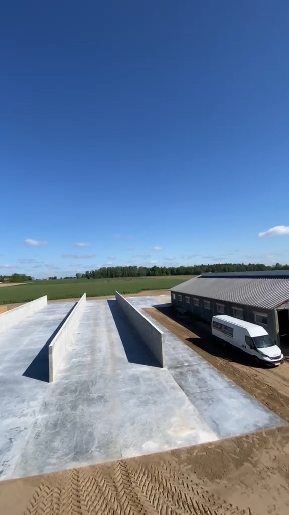 Betonowa konstrukcja z wysokimi ścianami, prawdopodobnie fundamenty pod budynek gospodarczy, z widocznym budynkiem w tle i zaparkowanym białym busem.