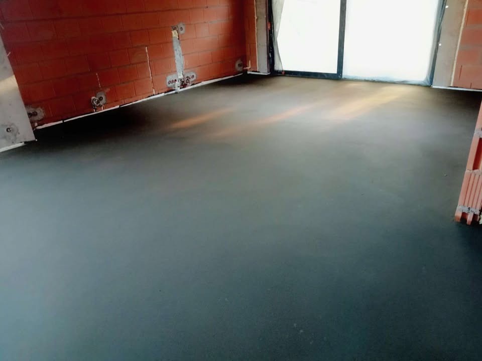 Gładka, ciemna posadzka w surowym wnętrzu z ceglanymi ścianami i dużym oknem balkonowym. Widoczne ślady po instalacjach na ścianach.