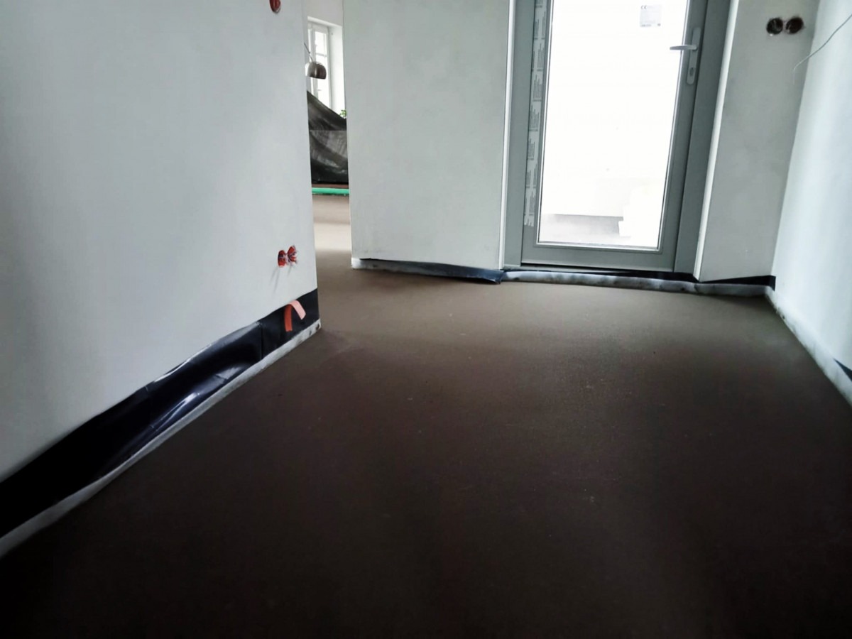 Wnętrze w trakcie prac wykończeniowych: wylewka podłogowa, folia ochronna przy ścianach, widoczne gniazdka elektryczne. Okno balkonowe w tle.