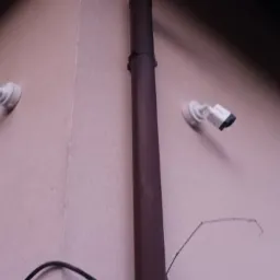 Dwie białe kamery monitoringu zamontowane na ścianie w rogu pomieszczenia, widoczne przewody i rura spustowa.