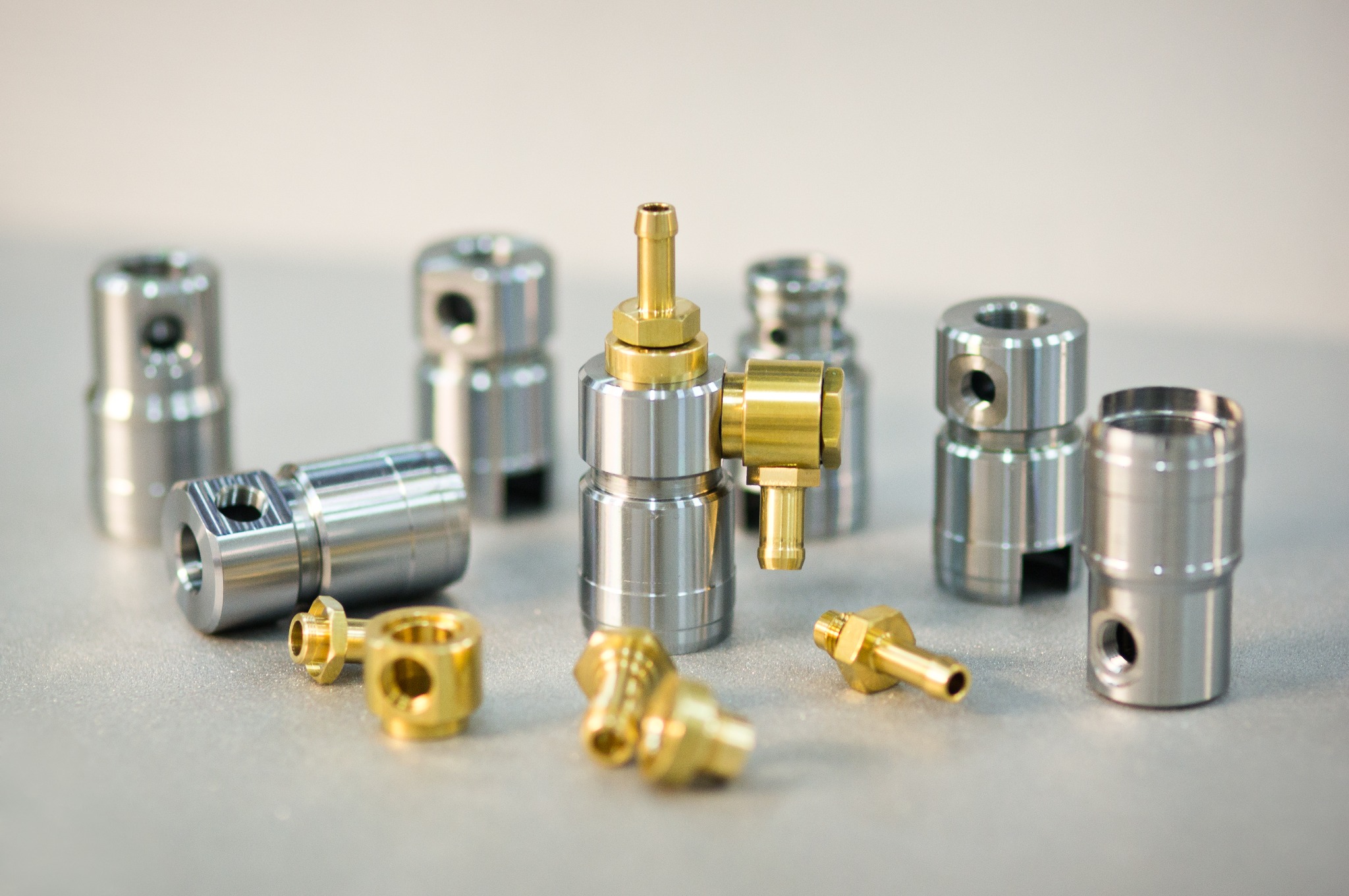 Precyzyjnie wykonane metalowe elementy o cylindrycznym kształcie, niektóre z widocznymi otworami i gwintami, w kolorze srebrnym i złotym, rozłożone na szarym tle.