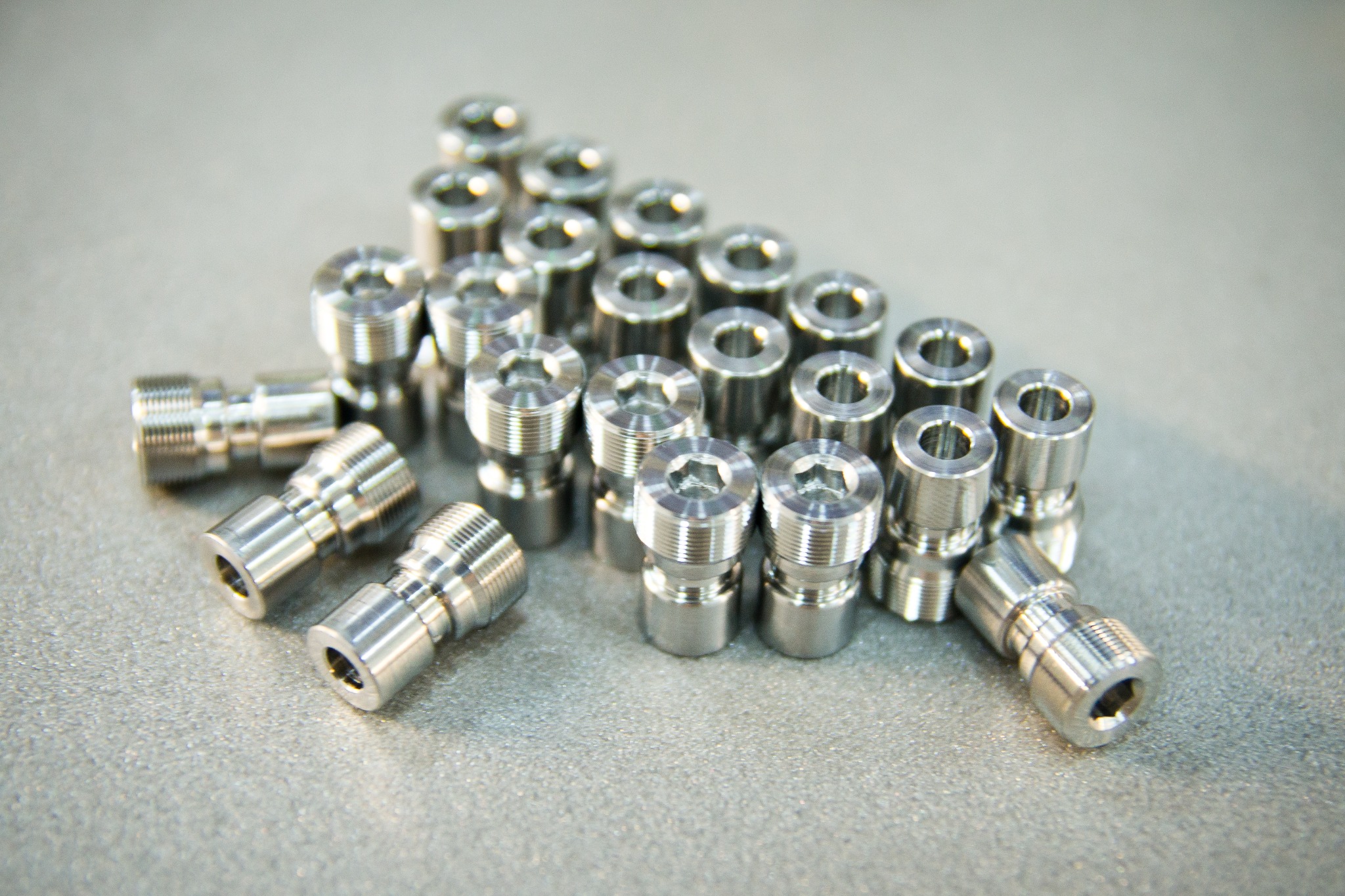 Partia precyzyjnie obrobionych, metalowych elementów z gwintem i otworem, ułożonych w formację trójkąta na szarym tle.