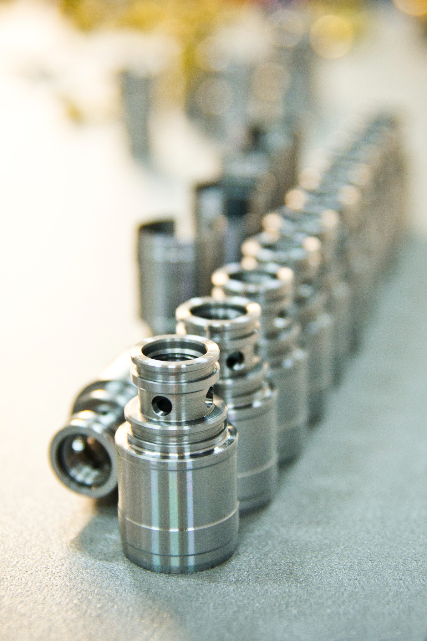 Rząd precyzyjnie wykonanych, metalowych elementów cylindrycznych z otworami, ułożonych na szarej powierzchni, z widocznymi śladami obróbki skrawaniem i delikatnym połyskiem.