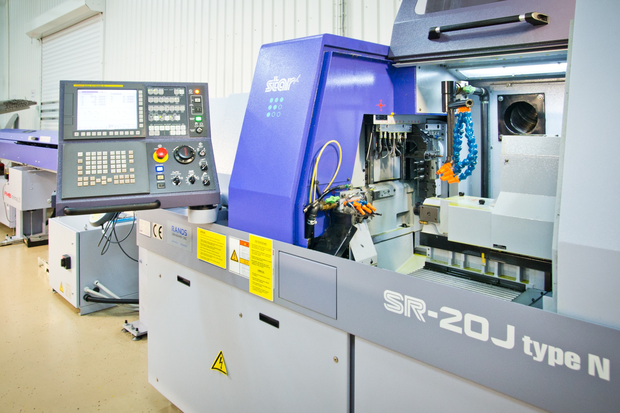 Szczegółowe ujęcie nowoczesnej tokarki CNC Star SR-20J type N z otwartą obudową, widoczne elementy robocze, panel sterowania z wyświetlaczem i przyciskami oraz ostrzeżenia bezpieczeństwa.