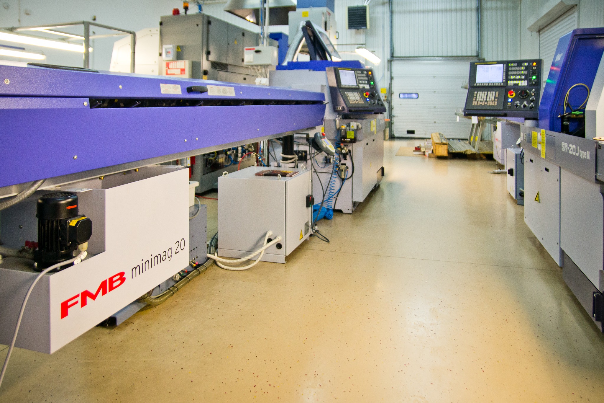 Widok hali produkcyjnej z kilkoma automatami tokarskimi CNC, z widocznym panelem sterowania, podajnikiem prętów FMB minimag 20, oraz bramą wjazdową w tle.
