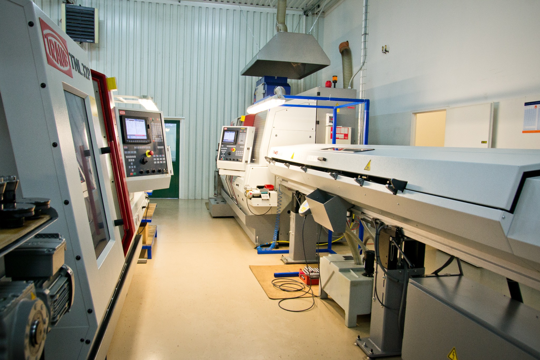 Widok na halę produkcyjną z maszynami sterowanymi numerycznie, widoczne panele sterowania i osłony maszyn, podłoga wyłożona wykładziną przemysłową.