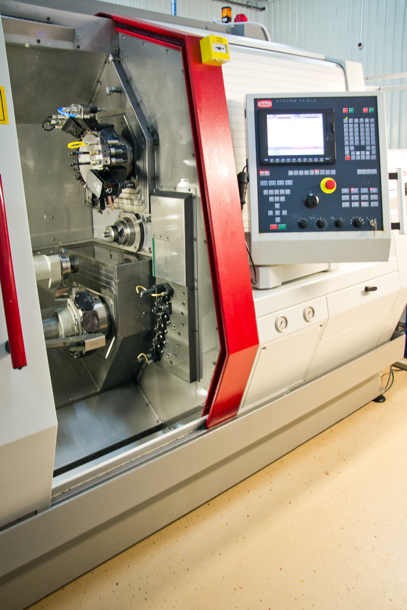 Precyzyjna obrabiarka CNC marki Traub z widocznym panelem sterowania i elementami obróbczymi w komorze roboczej, oświetlona światłem lampy, na podłodze z drobnymi plamkami.