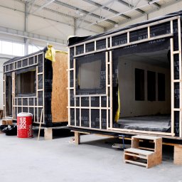 Izumrud Bud-Invest Sp. z o.o. - Dwa modułowe domy w trakcie budowy w hali, widoczne drewniane ramy, czarna membrana wiatroizolacyjna, płyty OSB, ocieplenie Rockwool, schodki wejściowe.