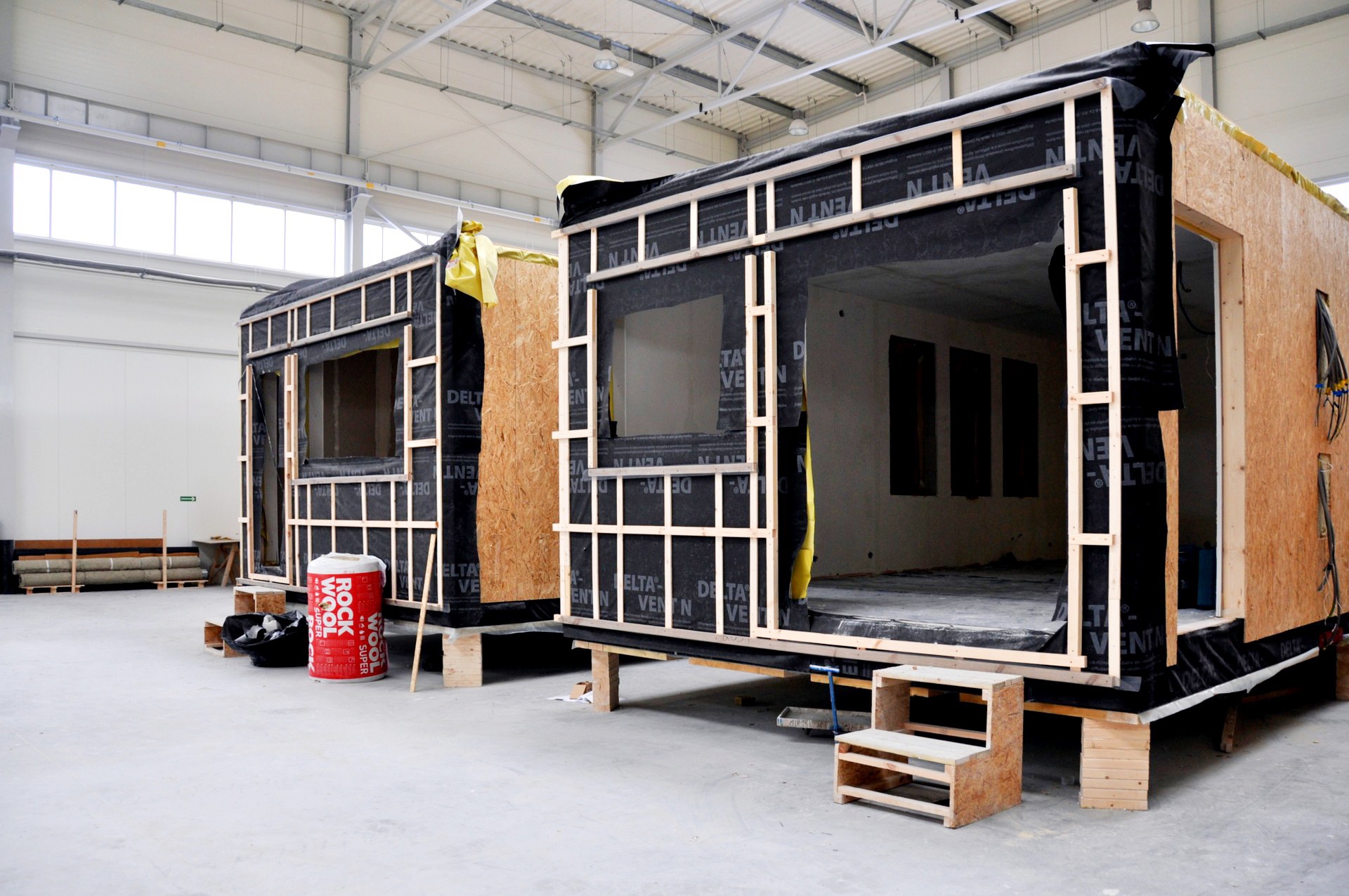 Dwa modułowe domy w trakcie budowy w hali, widoczne drewniane ramy, czarna membrana wiatroizolacyjna, płyty OSB, ocieplenie Rockwool, schodki wejściowe.