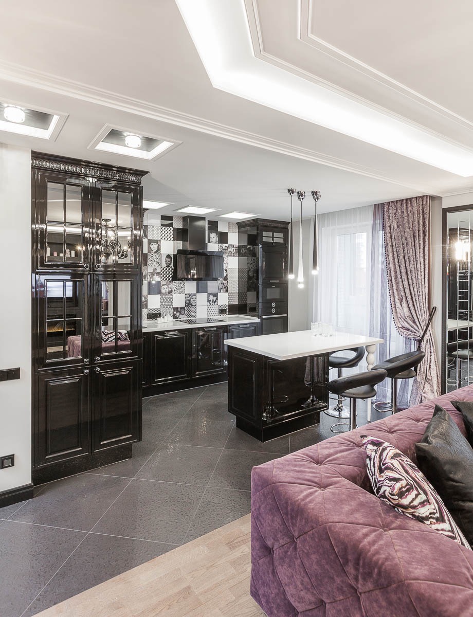 Eleganckie wnętrze: czarna zabudowa kuchenna z wyspą, patchworkowy backsplash, ozdobne oświetlenie sufitowe i fioletowa sofa w stylu glamour.