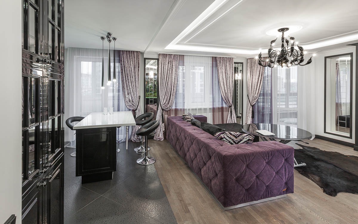 Eleganckie wnętrze salonu z fioletową sofą Chesterfield, czarnym barem z białym blatem, designerskim żyrandolem i zasłonami w odcieniach fioletu i bieli.