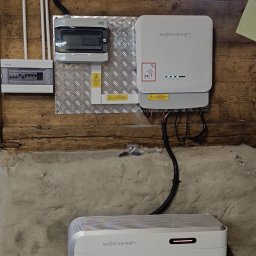 Anna Łukaszuk - Instalacja magazynu energii Sigenenergy w pomieszczeniu o drewnianych ścianach i betonowej podmurówce. Widoczne skrzynki elektryczne i okno z widokiem na drzewa.