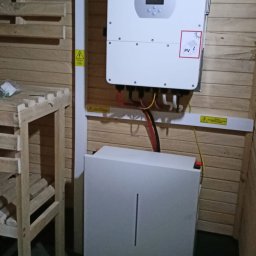 Anna Łukaszuk - Zamontowany inwerter Deye z podłączonymi kablami i akumulatorem na tle drewnianej ściany. Widoczna skrzynka z bezpiecznikami i ostrzeżenia o napięciu.