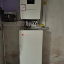 Anna Łukaszuk - Biały inwerter z etykietą PV i magazyn energii Felicitysolar na tle ściany. Widoczne kable i skrzynka elektryczna. Instalacja w pomieszczeniu o surowym wykończeniu.