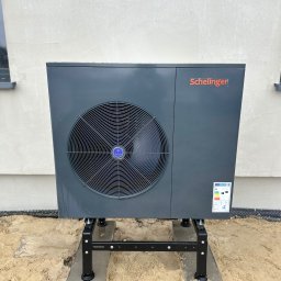 Anna Łukaszuk - Szara pompa ciepła Schelinger na czarnym stelażu, ustawiona przy jasnej ścianie budynku z widocznym oknem. Widoczna etykieta energetyczna na urządzeniu.