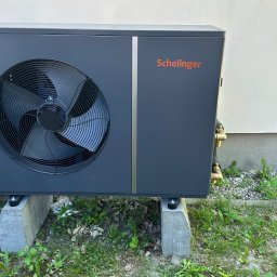 Anna Łukaszuk - Szara pompa ciepła Schelinger na betonowych podstawach, zainstalowana przy ścianie budynku. Widoczne przyłącza i wentylator. Trawa i kamienie wokół.