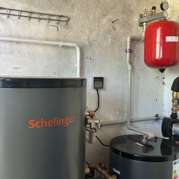 Anna Łukaszuk - Szary zbiornik Schelinger i czarny zbiornik z pompą obiegową w kotłowni. Widoczne rury, zawory i manometr. Tło stanowi surowa, betonowa ściana.