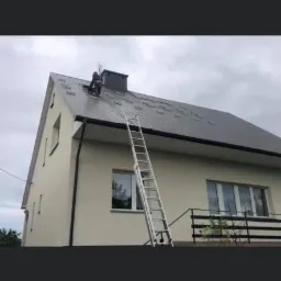 Pracownik na dachu domu jednorodzinnego z blachy trapezowej, podczas montażu lub naprawy. Aluminiowa drabina oparta o ścianę. Widoczne okna i orynnowanie.