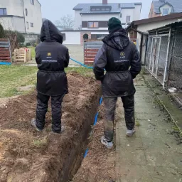 Dwaj pracownicy w odzieży roboczej z logo firmy stoją nad wykopem, w którym ułożona jest niebieska rura. W tle zabudowa jednorodzinna i pochmurne niebo.