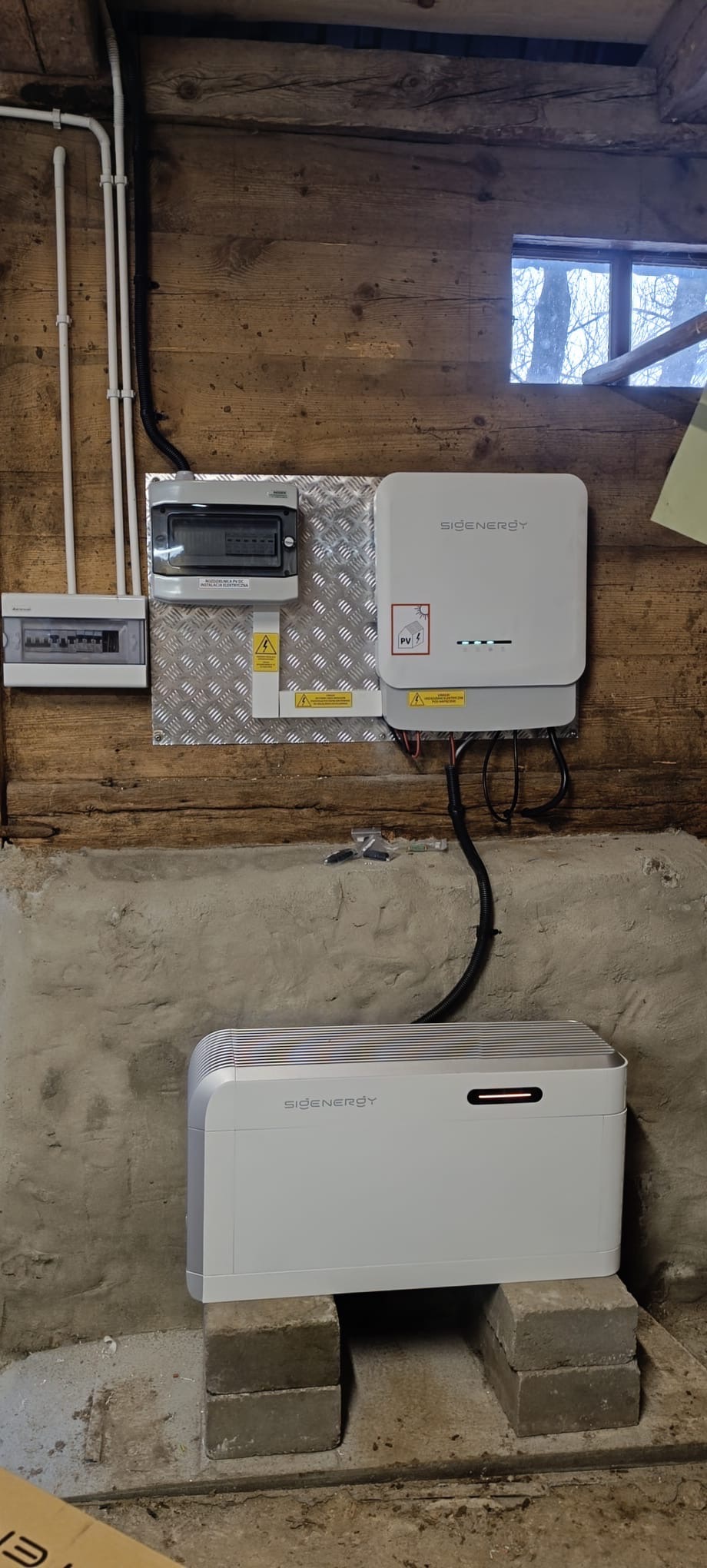 Instalacja magazynu energii Sigenenergy w pomieszczeniu o drewnianych ścianach i betonowej podmurówce. Widoczne skrzynki elektryczne i okno z widokiem na drzewa.