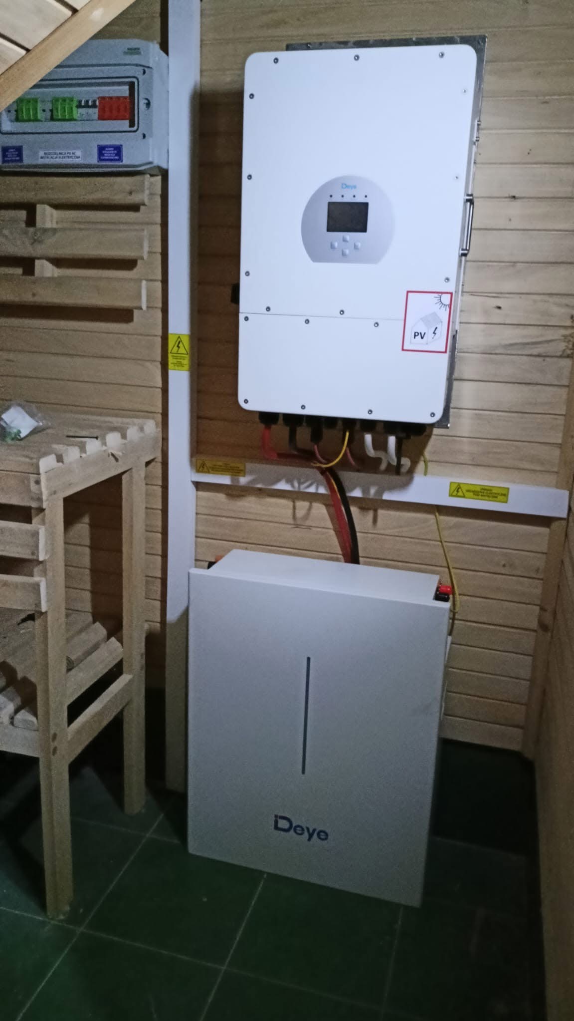Zamontowany inwerter Deye z podłączonymi kablami i akumulatorem na tle drewnianej ściany. Widoczna skrzynka z bezpiecznikami i ostrzeżenia o napięciu.
