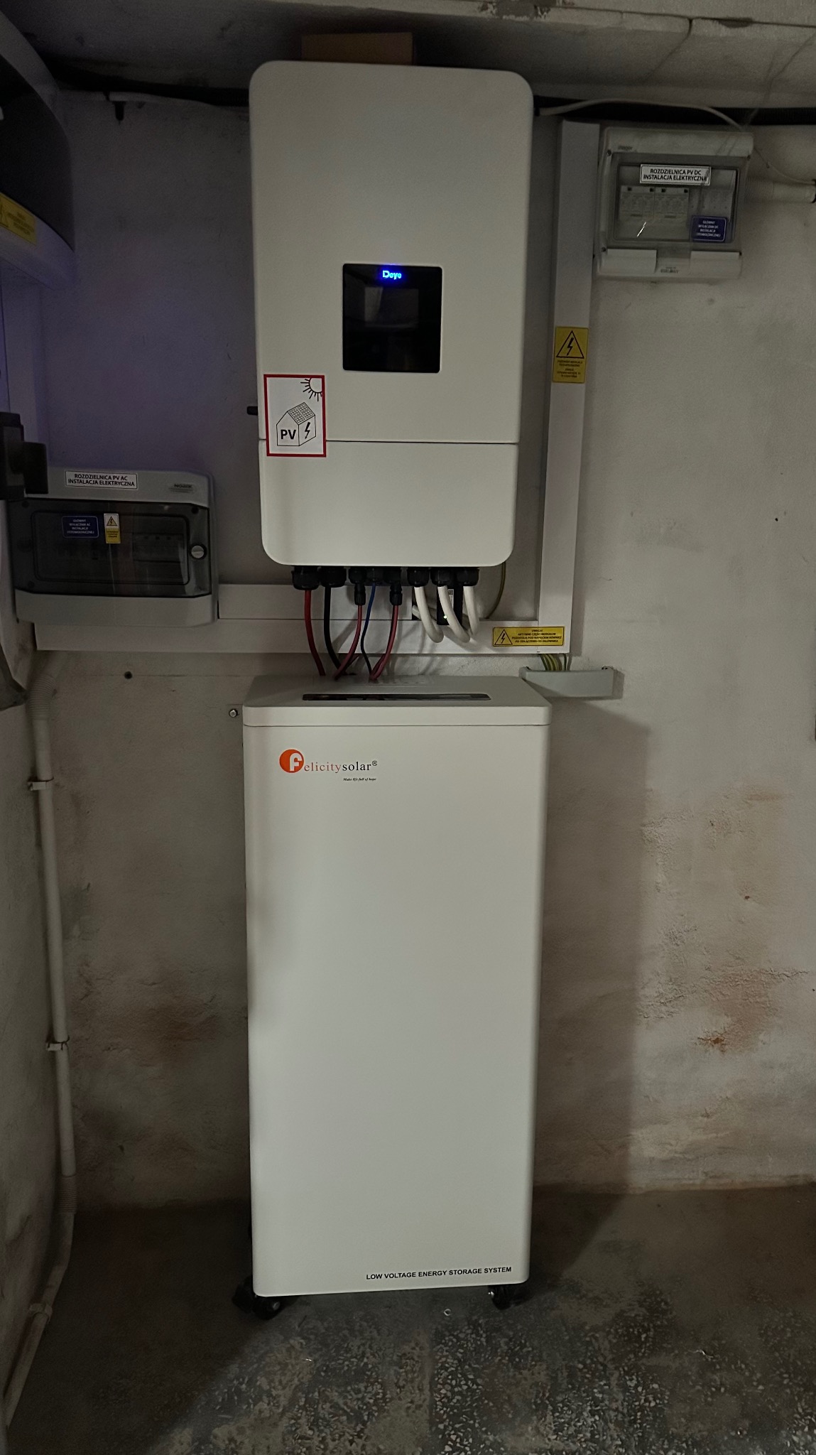Biały inwerter z etykietą PV i magazyn energii Felicitysolar na tle ściany. Widoczne kable i skrzynka elektryczna. Instalacja w pomieszczeniu o surowym wykończeniu.