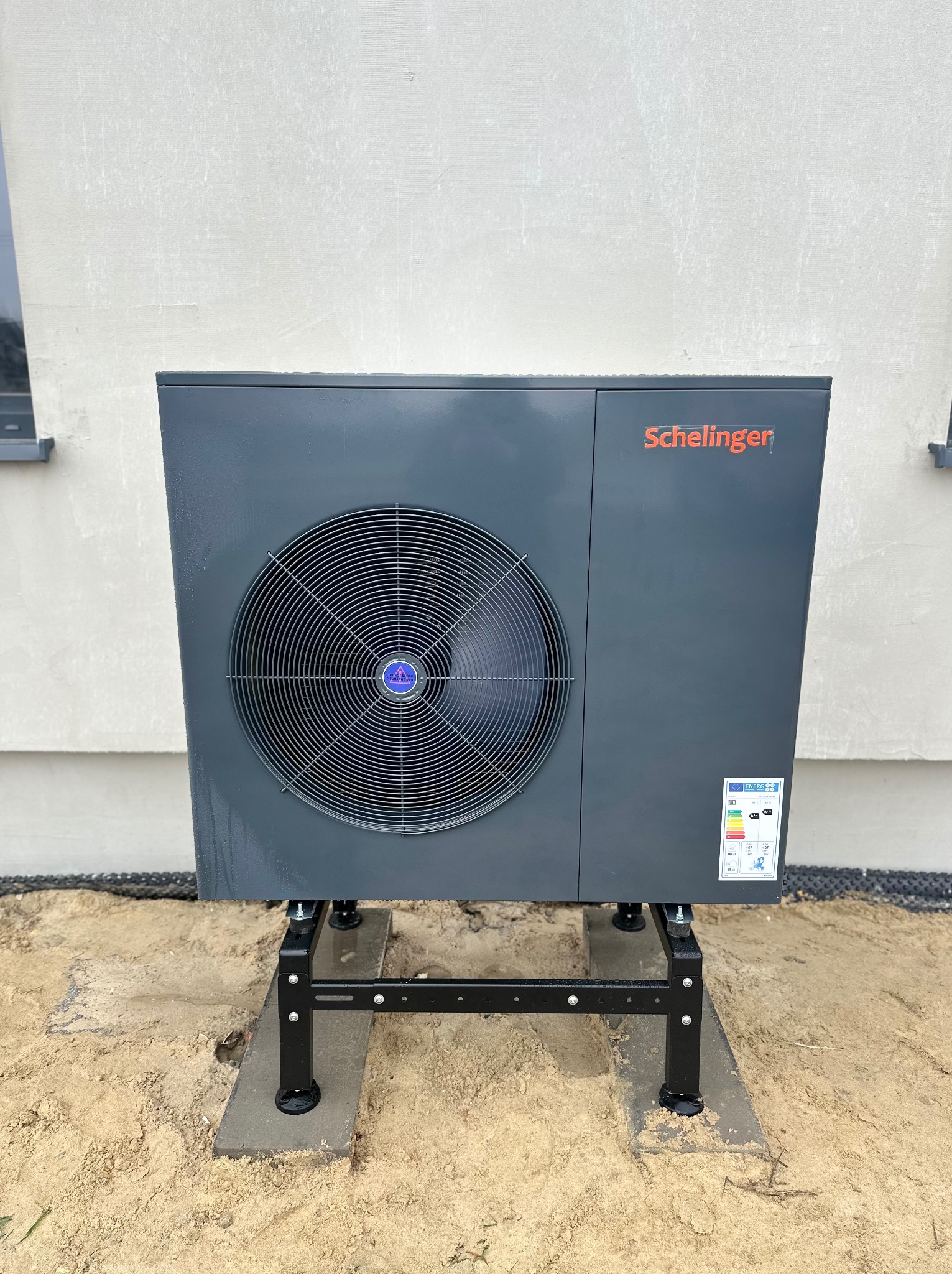 Szara pompa ciepła Schelinger na czarnym stelażu, ustawiona przy jasnej ścianie budynku z widocznym oknem. Widoczna etykieta energetyczna na urządzeniu.