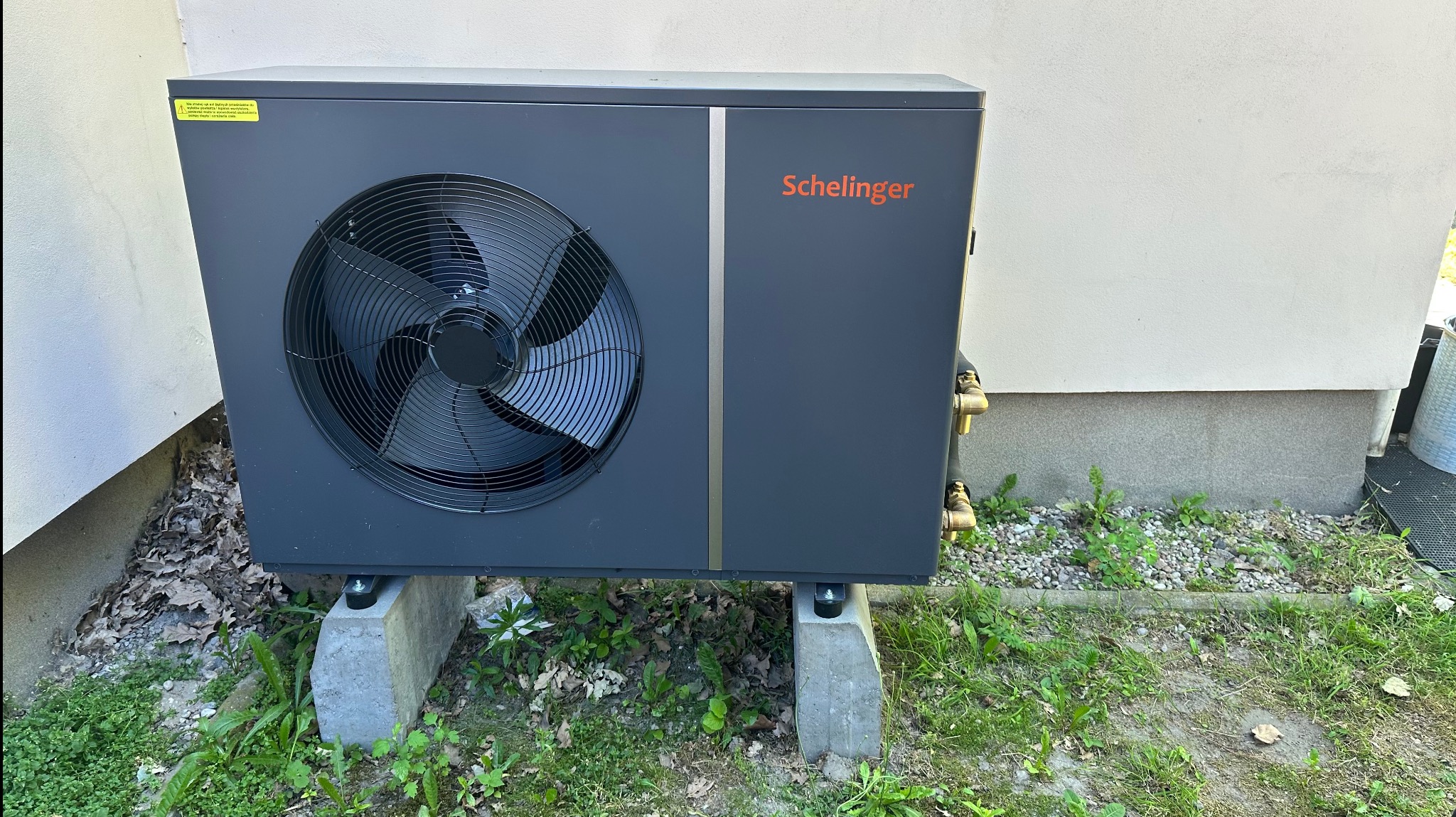 Szara pompa ciepła Schelinger na betonowych podstawach, zainstalowana przy ścianie budynku. Widoczne przyłącza i wentylator. Trawa i kamienie wokół.