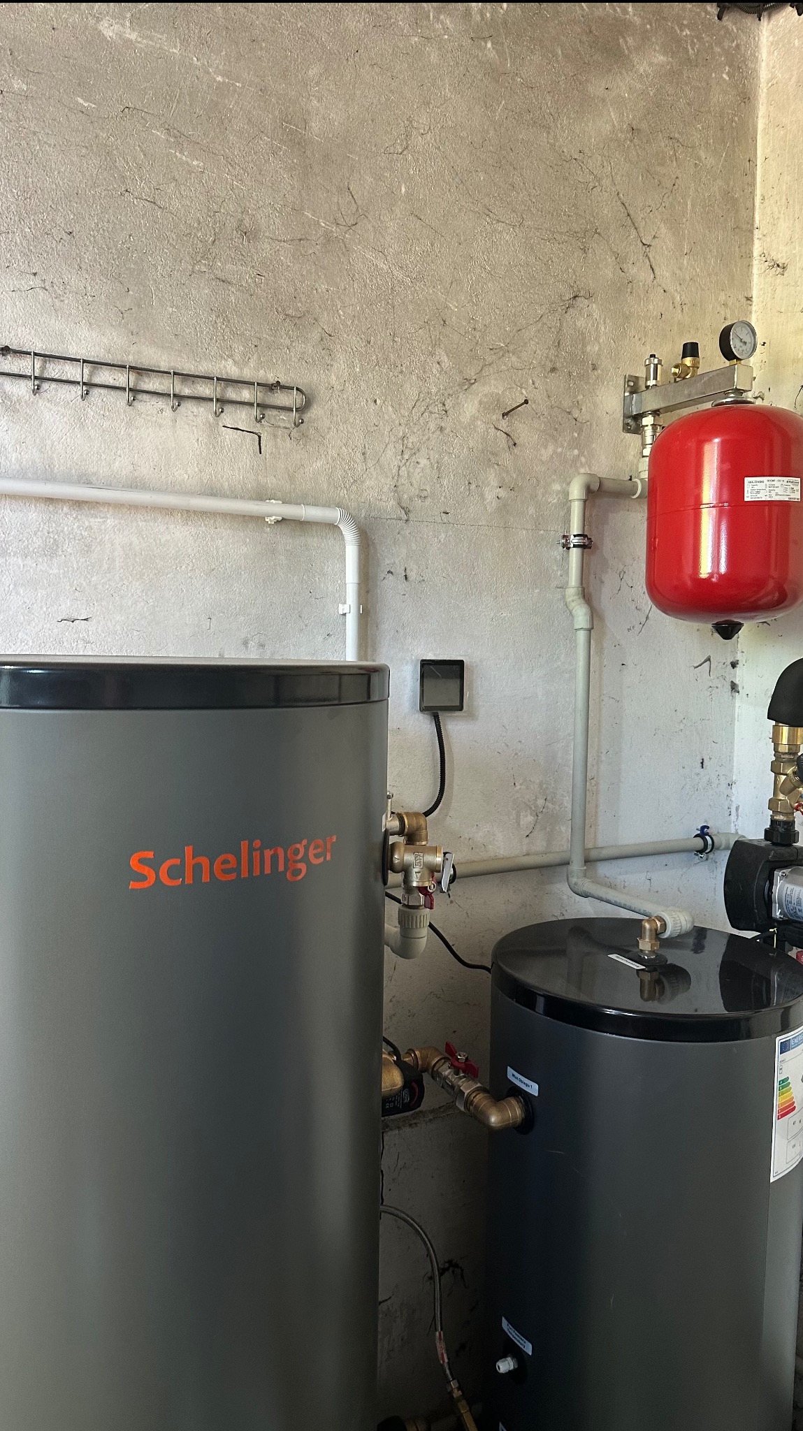 Szary zbiornik Schelinger i czarny zbiornik z pompą obiegową w kotłowni. Widoczne rury, zawory i manometr. Tło stanowi surowa, betonowa ściana.