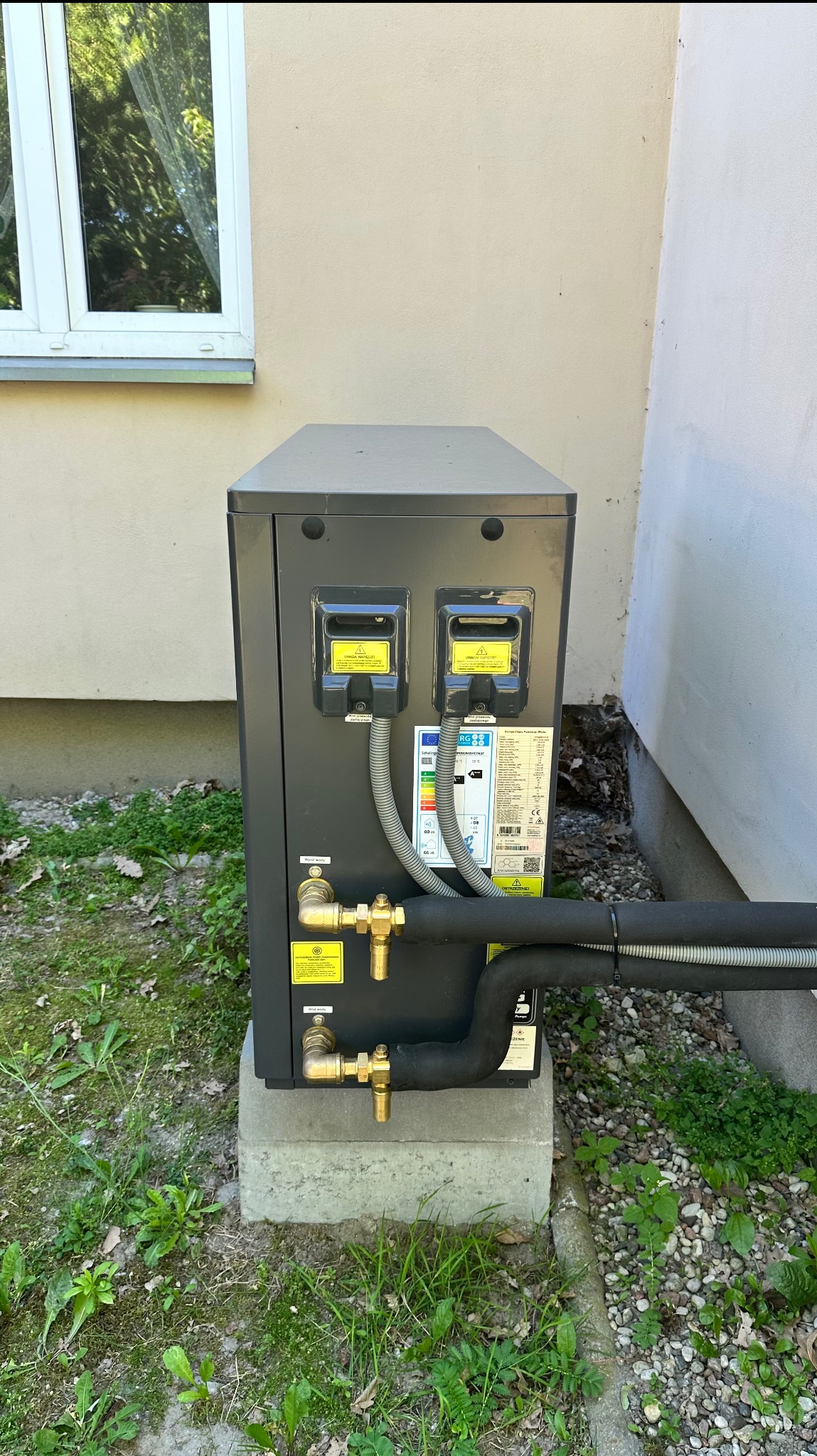 Zewnętrzna jednostka pompy ciepła na betonowym fundamencie, z widocznymi przyłączami i etykietami informacyjnymi, na tle ściany budynku i zieleni.