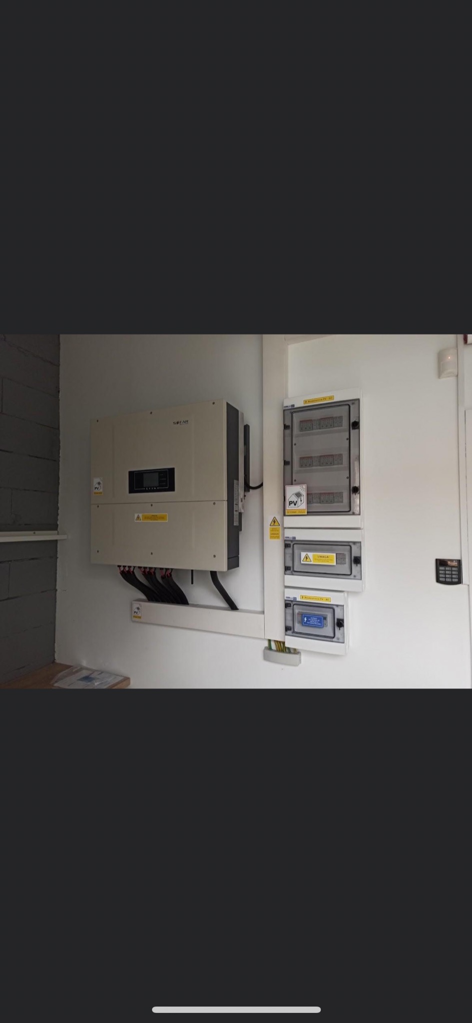 Zamontowany inwerter fotowoltaiczny marki SOFAR z podłączonymi kablami oraz rozdzielnica elektryczna z oznaczeniami ostrzegawczymi PV, zainstalowane na białej ścianie wewnątrz budynku.