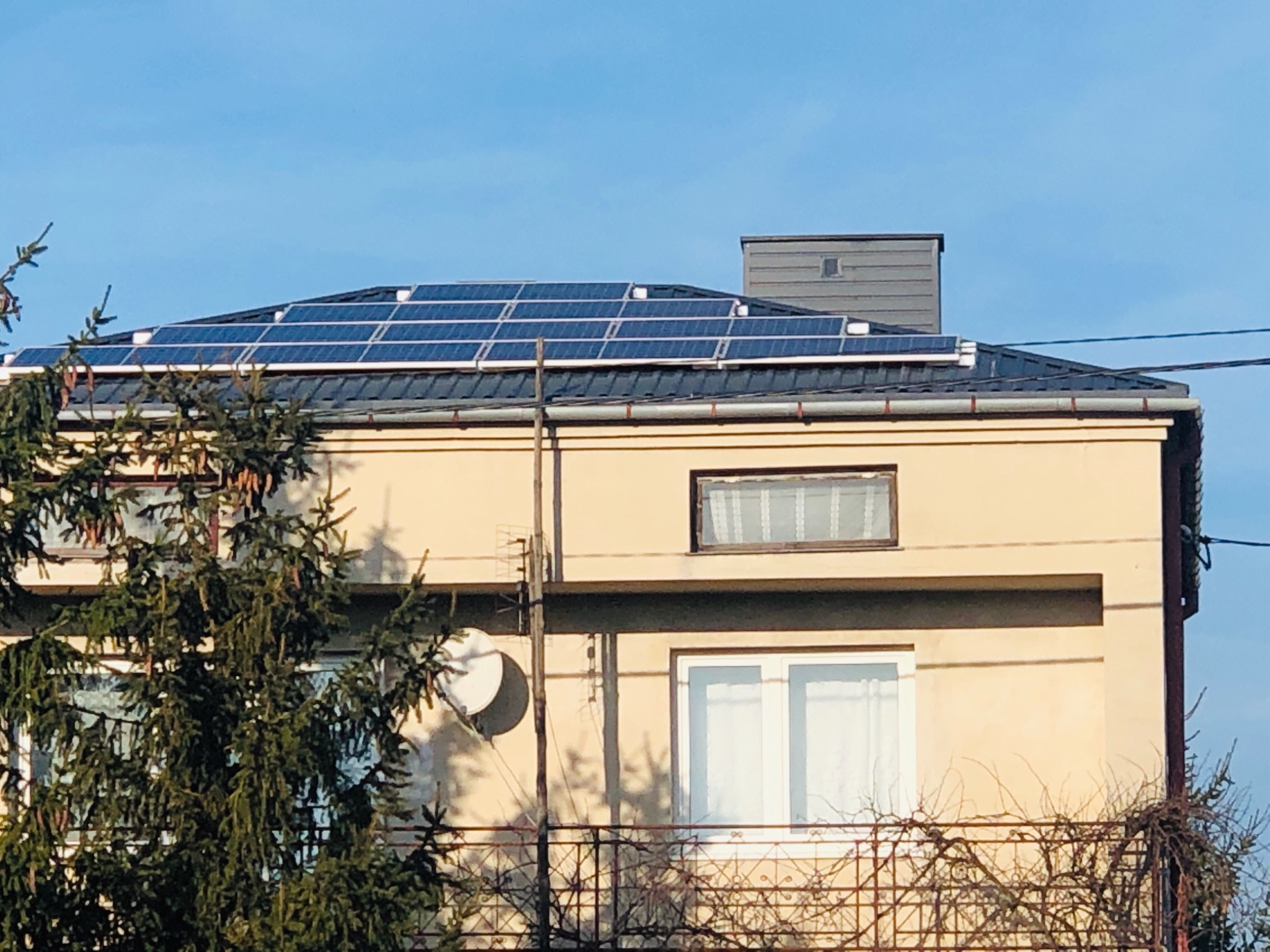 Panele słoneczne na dachu dwupiętrowego domu z beżową elewacją, widoczne z poziomu ulicy, z drzewem iglastym w pierwszym planie.