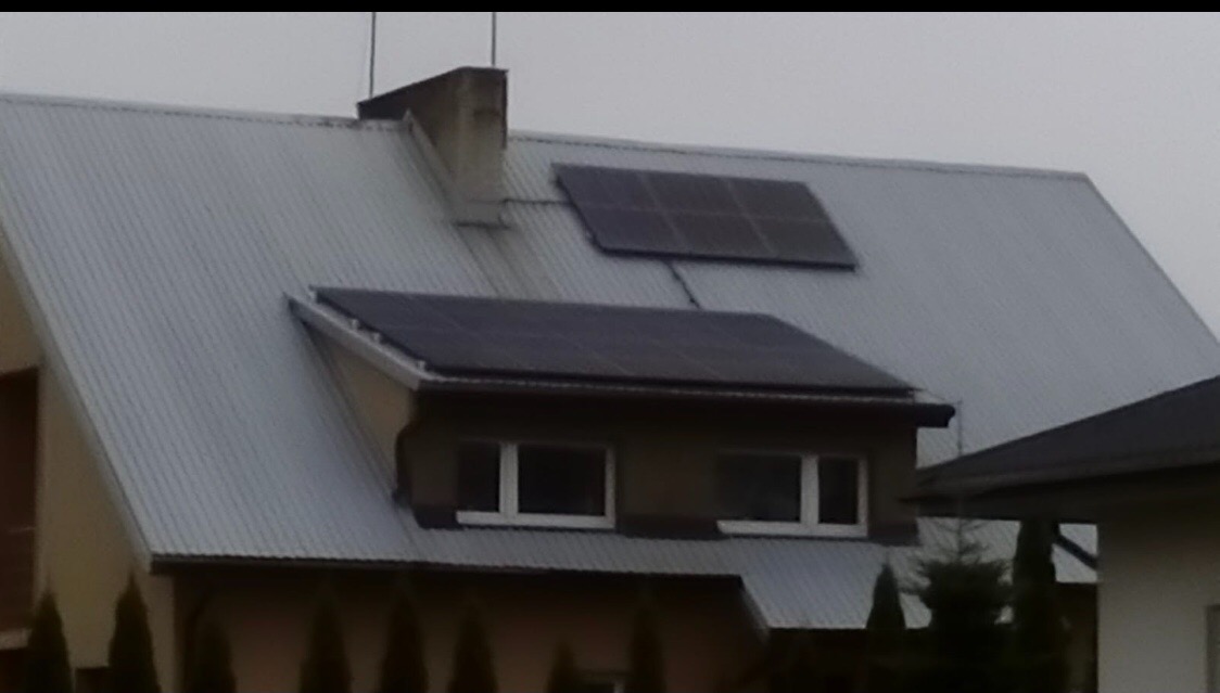 Dom jednorodzinny z szarego falistego metalu na dachu, na którym zamontowano dwa zestawy paneli słonecznych, jeden nad oknem mansardowym, drugi bliżej komina.