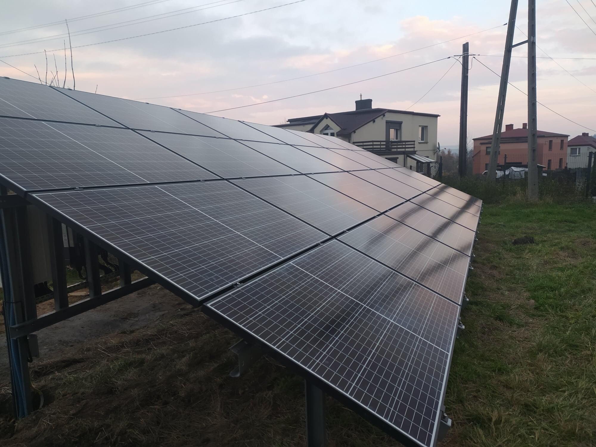Panel słoneczny zainstalowany na metalowej konstrukcji na trawiastym terenie, z domami i słupami energetycznymi w tle pod pochmurnym niebem.