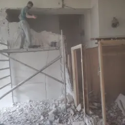 Pracownik na rusztowaniu skuwa resztki tynku ze ściany, widoczne gruz i narzędzia, w tle ościeżnice drzwiowe.