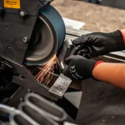 Operator w czarnych rękawicach ochronnych szlifuje metalowy element na szlifierce taśmowej, iskry lecą w kierunku operatora.