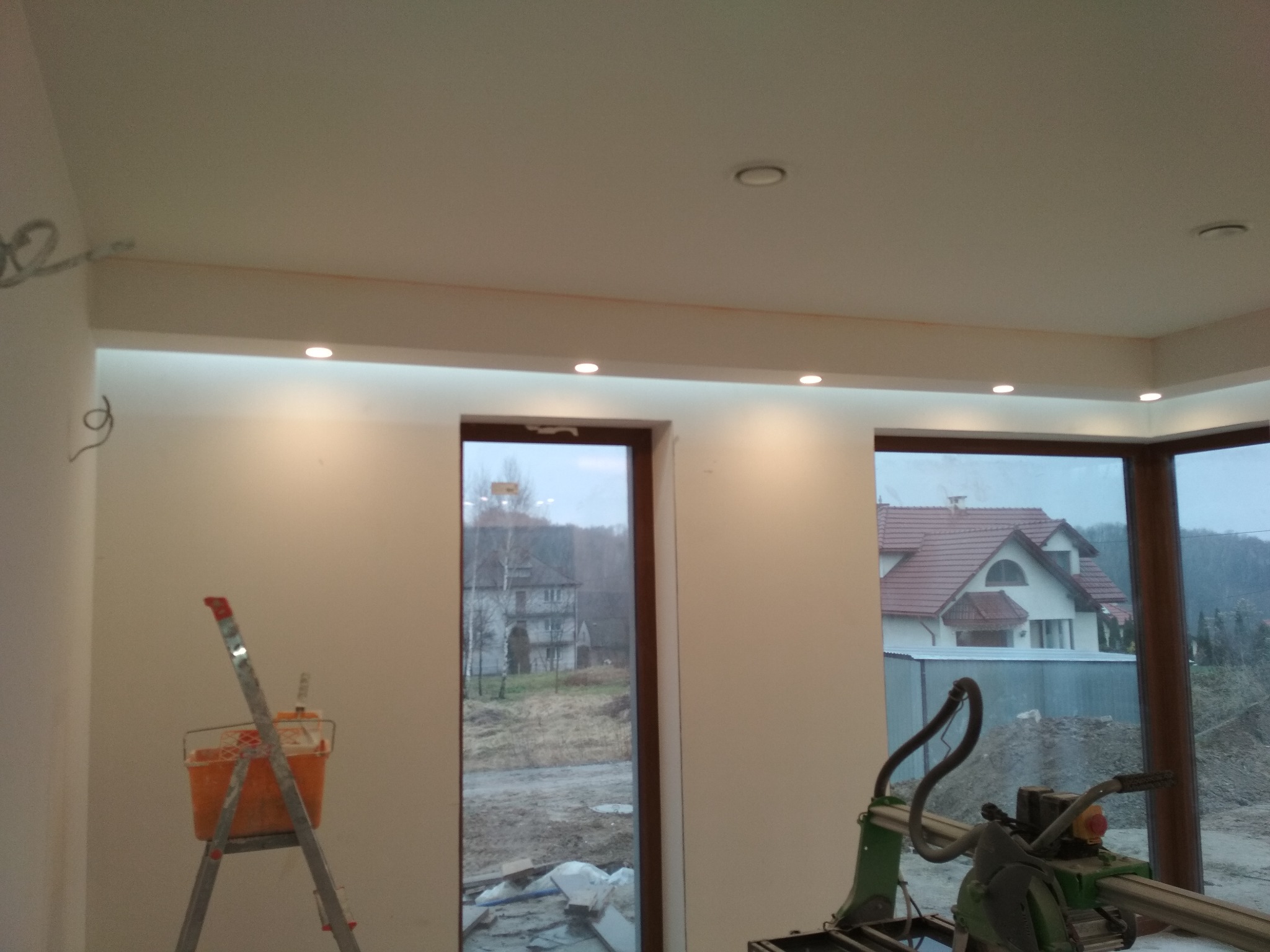 Wnętrze w trakcie remontu, widoczna drabina z pomarańczowym wiadrem, przecinarka do płytek przy oknie z widokiem na dom, sufit z oświetleniem punktowym i wystającymi kablami.
